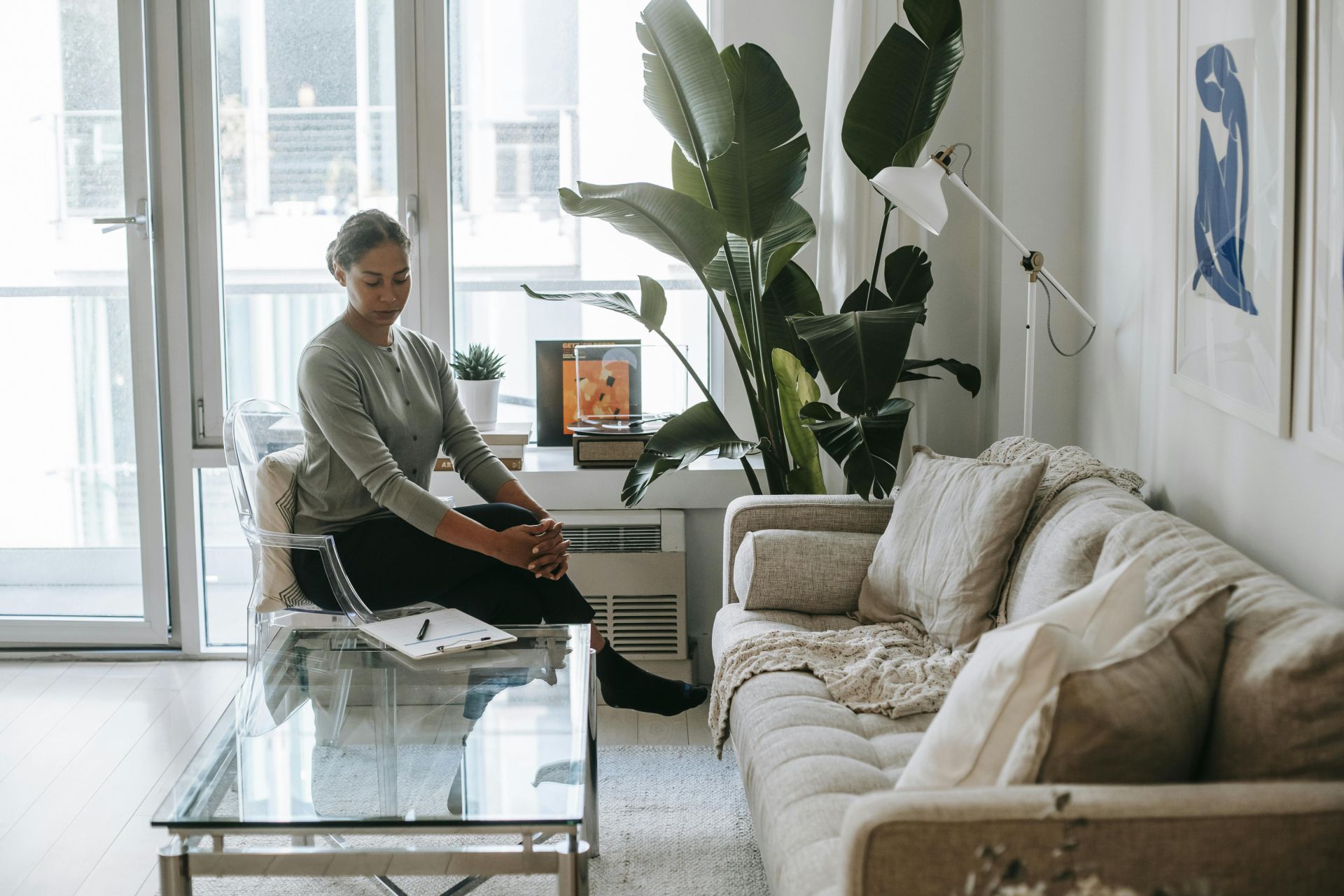 Peaceful ethnic female psychologist with crossed legs looking down while sitting near table with clipboard in modern office while waiting for patient